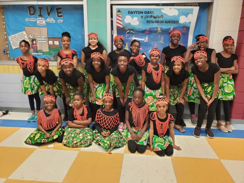 The photograph depicts a dance troupe wearing traditional clothing, posing in rows in front of a school bulletin board.