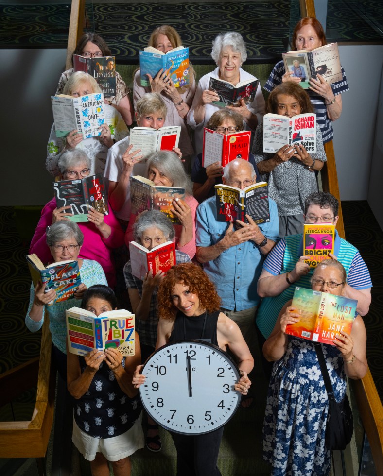 A group of folks from the Noontime Book Club stand on the steps at Central holding books in front of their faces, with one person holding a clock set to twelve o'clock.